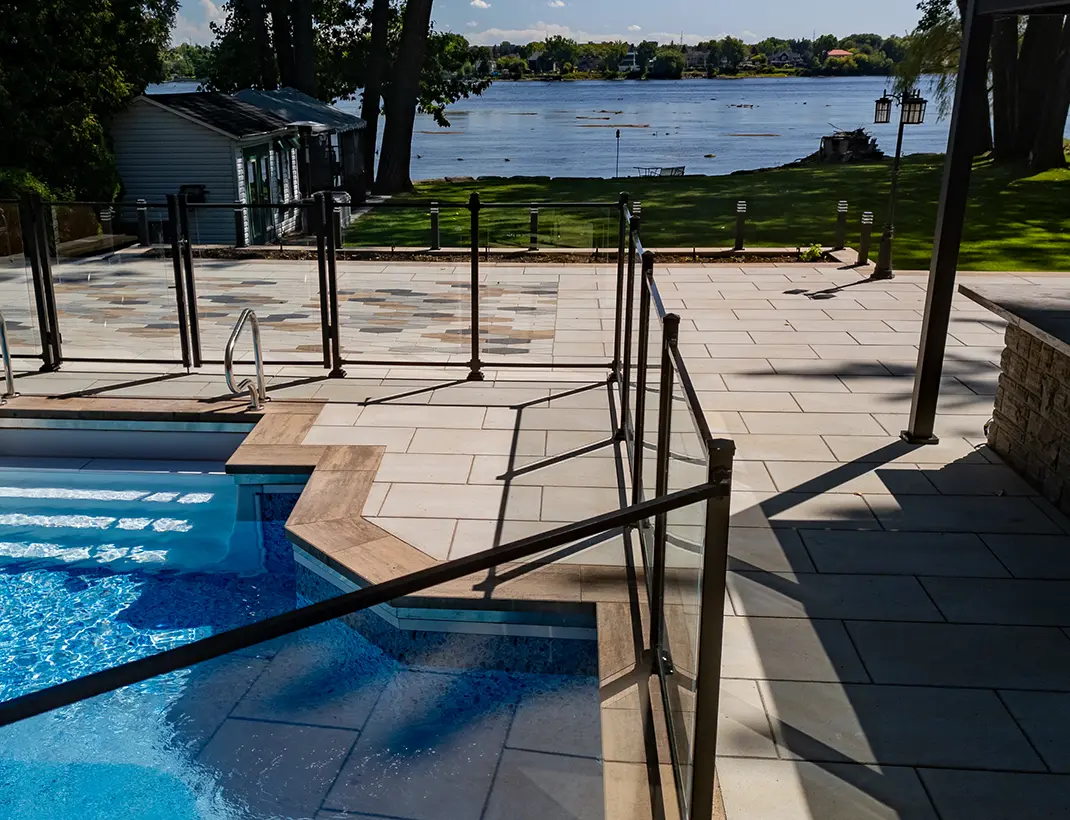 Terrasse de luxe en pavé uni entourant une piscine avec clôture de verre, offrant une vue panoramique sur un cours d'eau de la Rive-Sud.