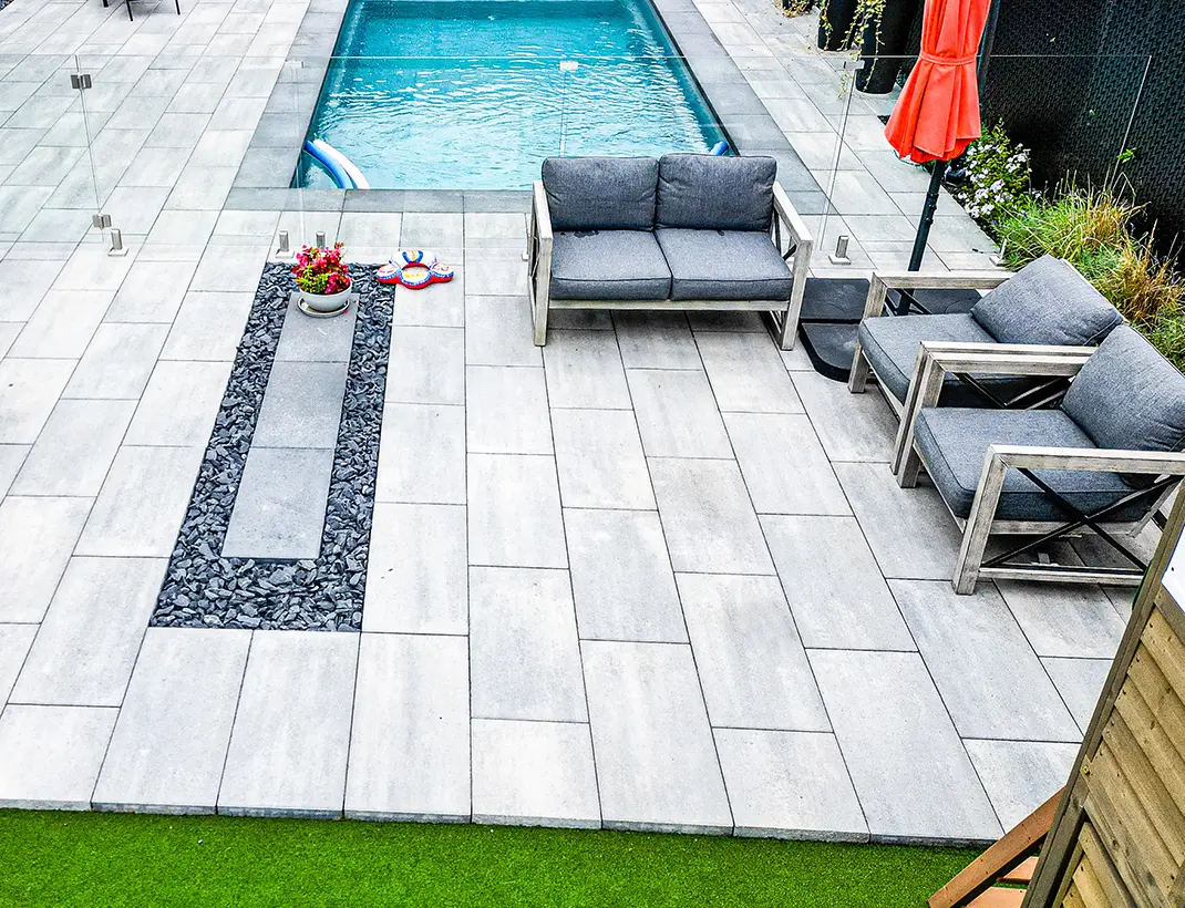 Aménagement d'une cour arrière avec dalles de pavé uni gris grand format, mobilier de jardin moderne et piscine creusée sécurisée par une paroi vitrée.